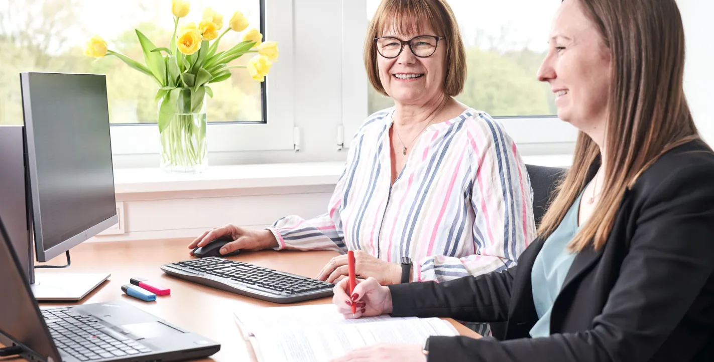 Zwei Kolleginnen arbeiten lächelnd an einem hellen Büro-Arbeitsplatz